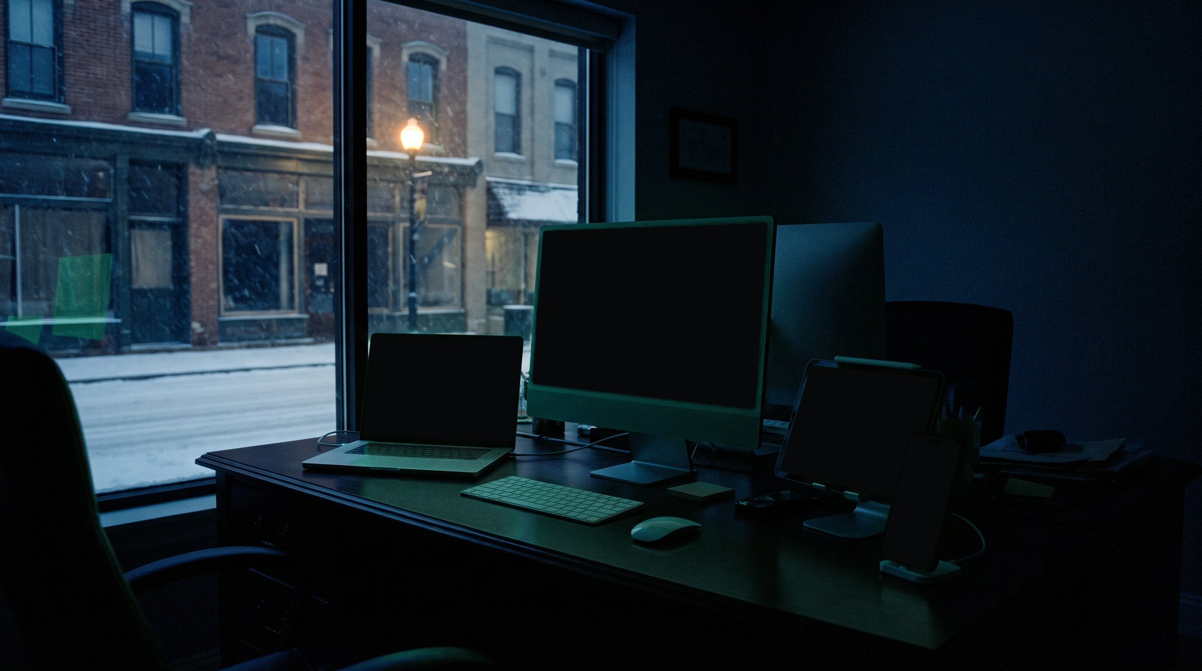 A dark office at night lit only by its screens: a phone showing a client website, a laptop with an AI concierge open, a tablet running a client platform and a monitor showing Orbit